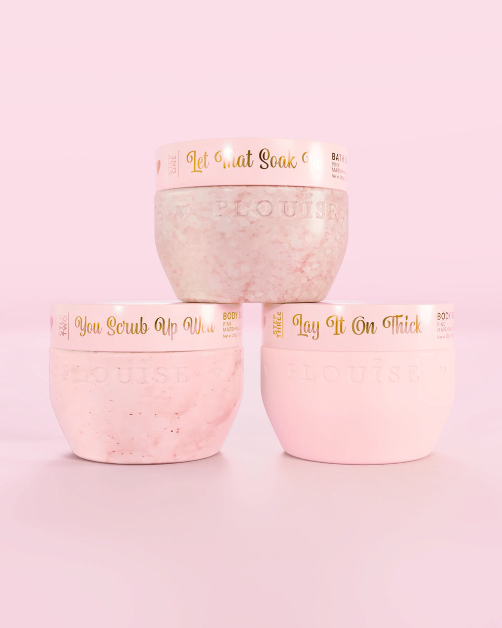 Tre contenitori rosa Marshmallow Hop Into Self-Care Pink di P.Louise con sali da bagno, scrub corpo e burro per il corpo con testo dorato su sfondo rosa