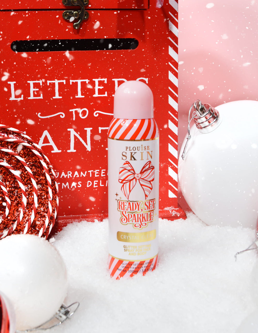 Una bottiglia di spray fissante P. Louise Ready Set Sparkle Christmas Glitter circondato da decorazioni festive come neve, palline e una cassetta delle lettere natalizie