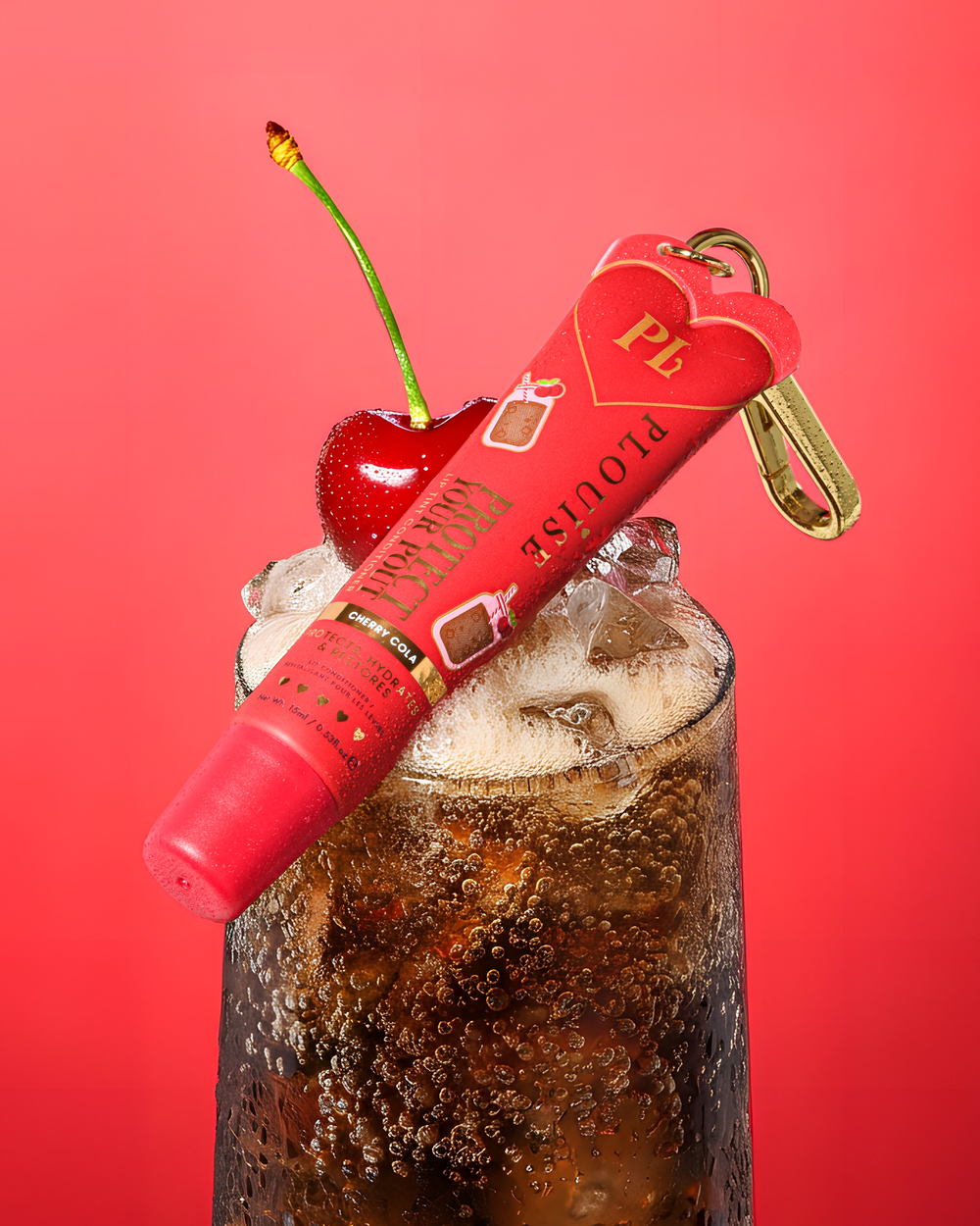 Balsamo per labbra colorato al profumo di cola e ciliegia rossa con clip dorata sopra un bicchiere di cola alla ciliegia con una ciliegia