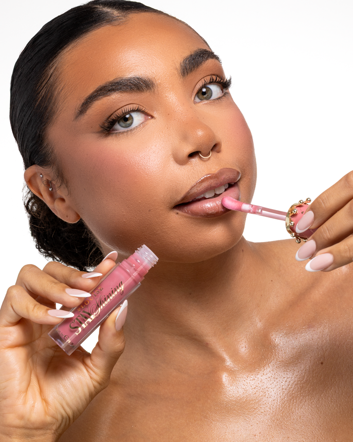 Un primo piano di una donna che applica un prodotto labbra liquido P.Louise Stay Shining Lip Oil rosa con un applicatore a forma di cerbiatto mentre tiene il tubetto nell'altra mano, su uno sfondo bianco pulito.