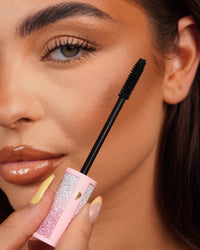 Primo piano di una bacchetta di mascara P.Louise Cry Baby tenuta vicino all'occhio di una persona con un tubetto di mascara rosa scintillante.