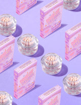 Contenitori trasparenti di P.Louise DOse of Dreams Viatmin C Capsule Face Cream con capsule rosa al loro interno circondate da scatole di cereali rosa su sfondo viola