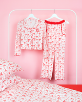 Set di pantaloni pigiama per ragazze P.Louise con fiocco natalizio caratterizzato da un disegno di fiocco rosso su tutta la superficie su strisce rosa e bianche appese alle grucce in una camera da letto rosa con biancheria da letto abbinata