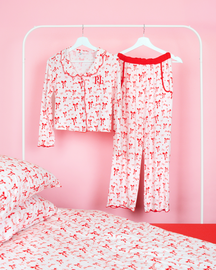 Set di pantaloni pigiama per ragazze P.Louise con fiocco natalizio caratterizzato da un disegno di fiocco rosso su tutta la superficie su strisce rosa e bianche appese alle grucce in una camera da letto rosa con biancheria da letto abbinata