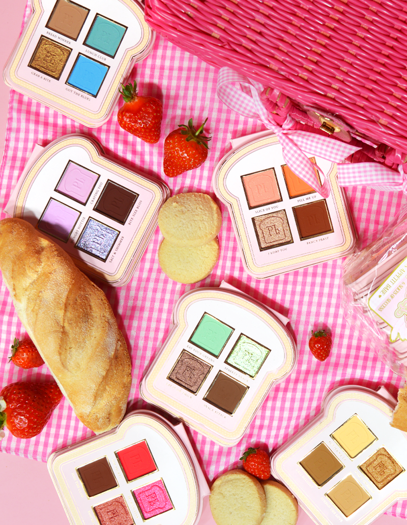 Un set di mini palette di ombretti in stile sandwich con varie sfumature di colori per il trucco visualizzate su una superficie a quadretti rosa, accompagnate da fragole e un pezzo di pane.
