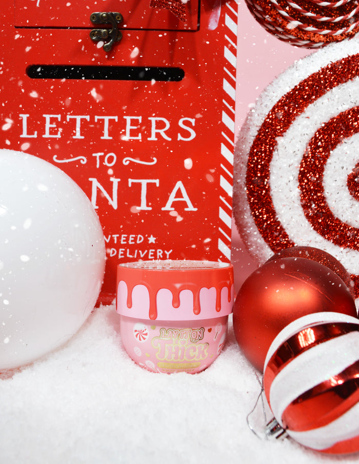 Un barattolo di P.Louise Lay It On Thick Body Butter nella variante Frosted Peppermint, esposto con decorazioni a tema natalizio attorno, comprese luci e regali di Natale.