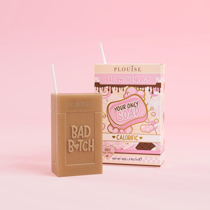 Una saponetta marrone con inciso "Bad Bitch", accompagnata da una confezione di succo colorato con l'etichetta "Calorific", presentata su uno sfondo rosa.