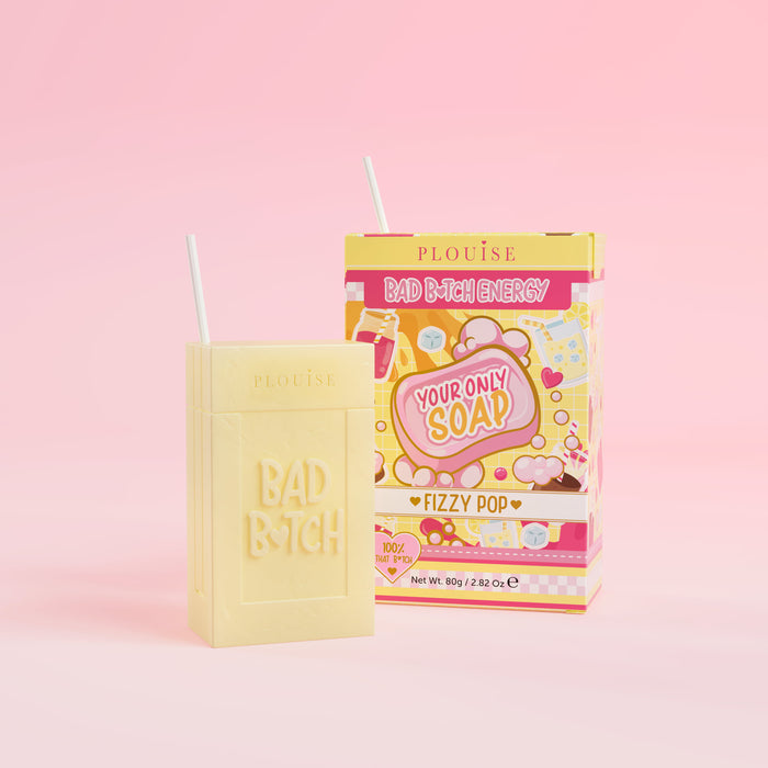 Una saponetta gialla con la scritta "Bad Bitch" incisa sopra, accompagnata da una confezione di succo colorato con l'etichetta "Fizzy Pop", presentata su uno sfondo rosa.
