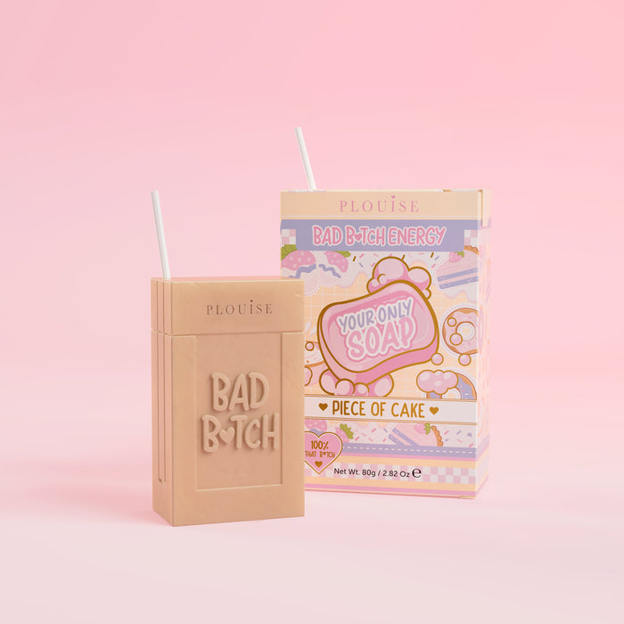 Una saponetta beige con la scritta "Bad Bitch" incisa sopra, accompagnata da una confezione di succo colorato con l'etichetta "Piece of Cake", presentata su uno sfondo rosa.