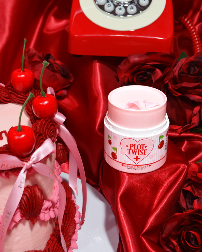 Un contenitore di P.Louise Plot Twist Make It Melt Makeup Cleansing Balm esposto su una superficie di tessuto rosso con elementi decorativi tra cui una ciliegia e un telefono rosso vintage sullo sfondo.