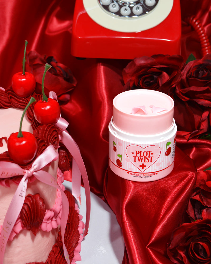 Un contenitore di P.Louise Plot Twist Make It Melt Makeup Cleansing Balm esposto su una superficie di tessuto rosso con elementi decorativi tra cui una ciliegia e un telefono rosso vintage sullo sfondo.