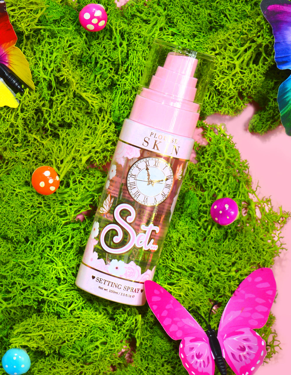 Una bottiglia di P. Louise Ready Set Slay Spray fissante a lunga durata circondato da elementi decorativi tra cui muschio finto colorato, farfalle e funghi.