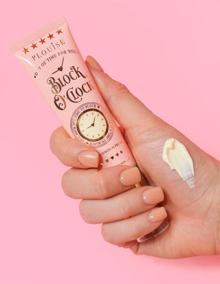 A hand holding a tube of P.Louise Block O'Clock Pore Perfecting Primer with product applied to the finger, on a pink background.